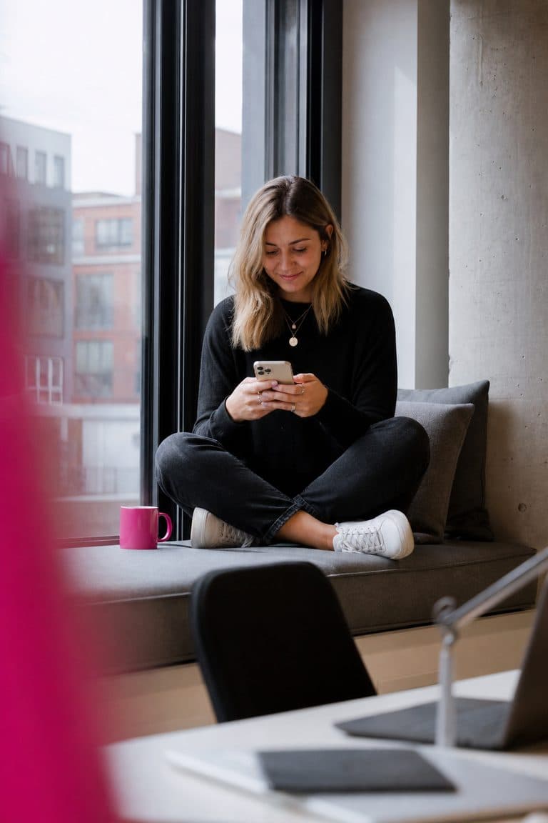 Community-Managerin beantwortet auf der Fensterbank im Büro Nachrichten auf dem Handy