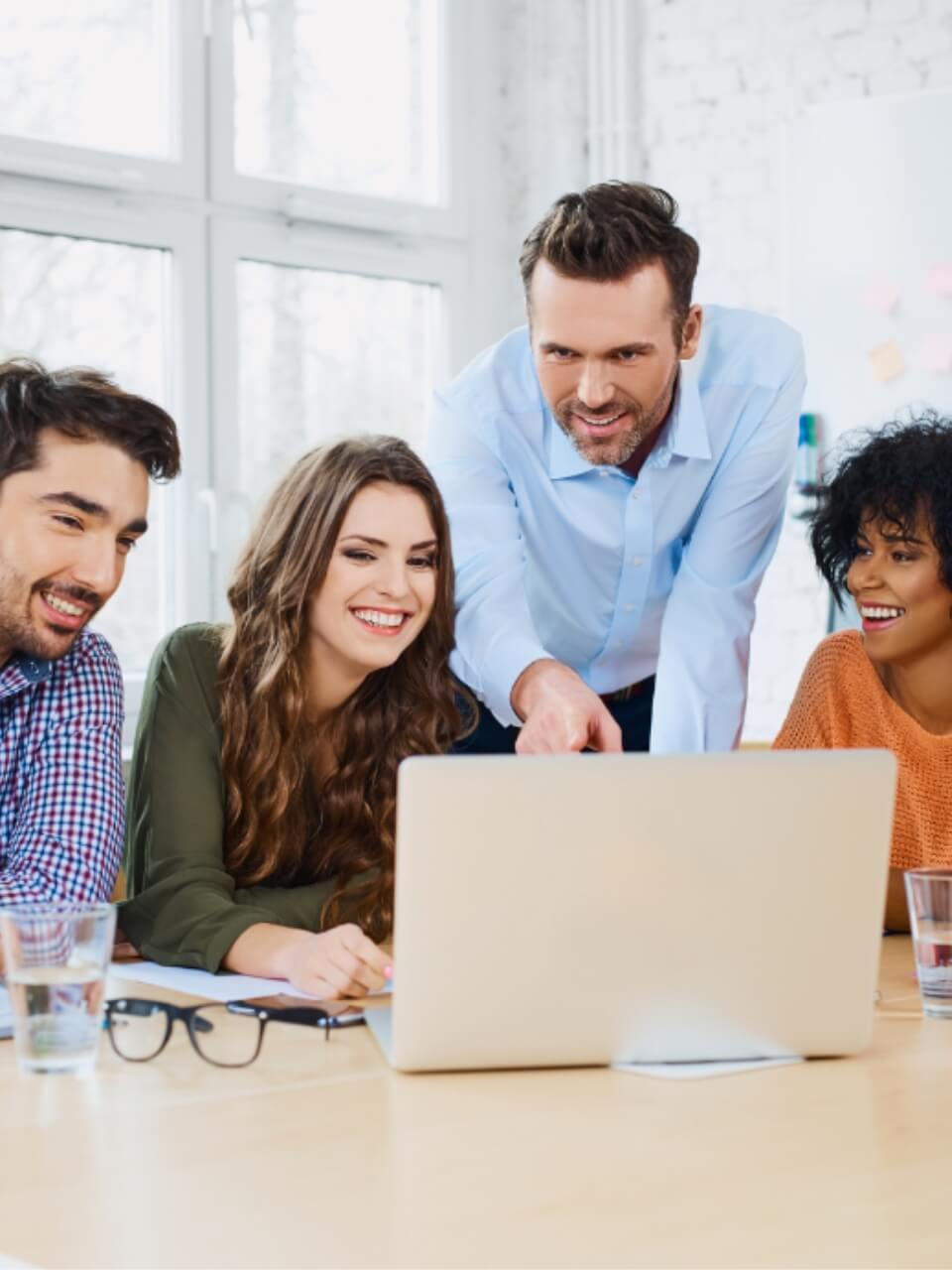 Vier lächelnde Kollegen betrachten gemeinsam einen Laptop auf einem hellen Bürotisch, offensichtlich in einer angenehmen Teamarbeitssituation.